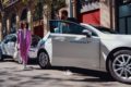 two white sharenow cars are parallel parked on side of the street. one driver is getting out of the car and greets another pedestrian.