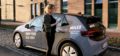 an electric miles car is parallel parked, a woman uses her smartphone to rent and unlock it. on the car a slogan says a