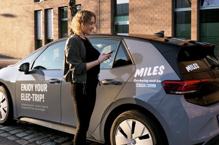 an electric miles car is parallel parked, a woman uses her smartphone to rent and unlock it. on the car a slogan says a